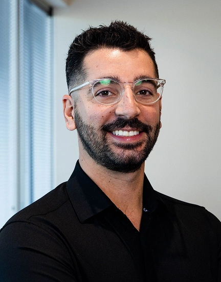 Professional headshot of a smiling man wearing glasses and dark shirt in bright office setting.