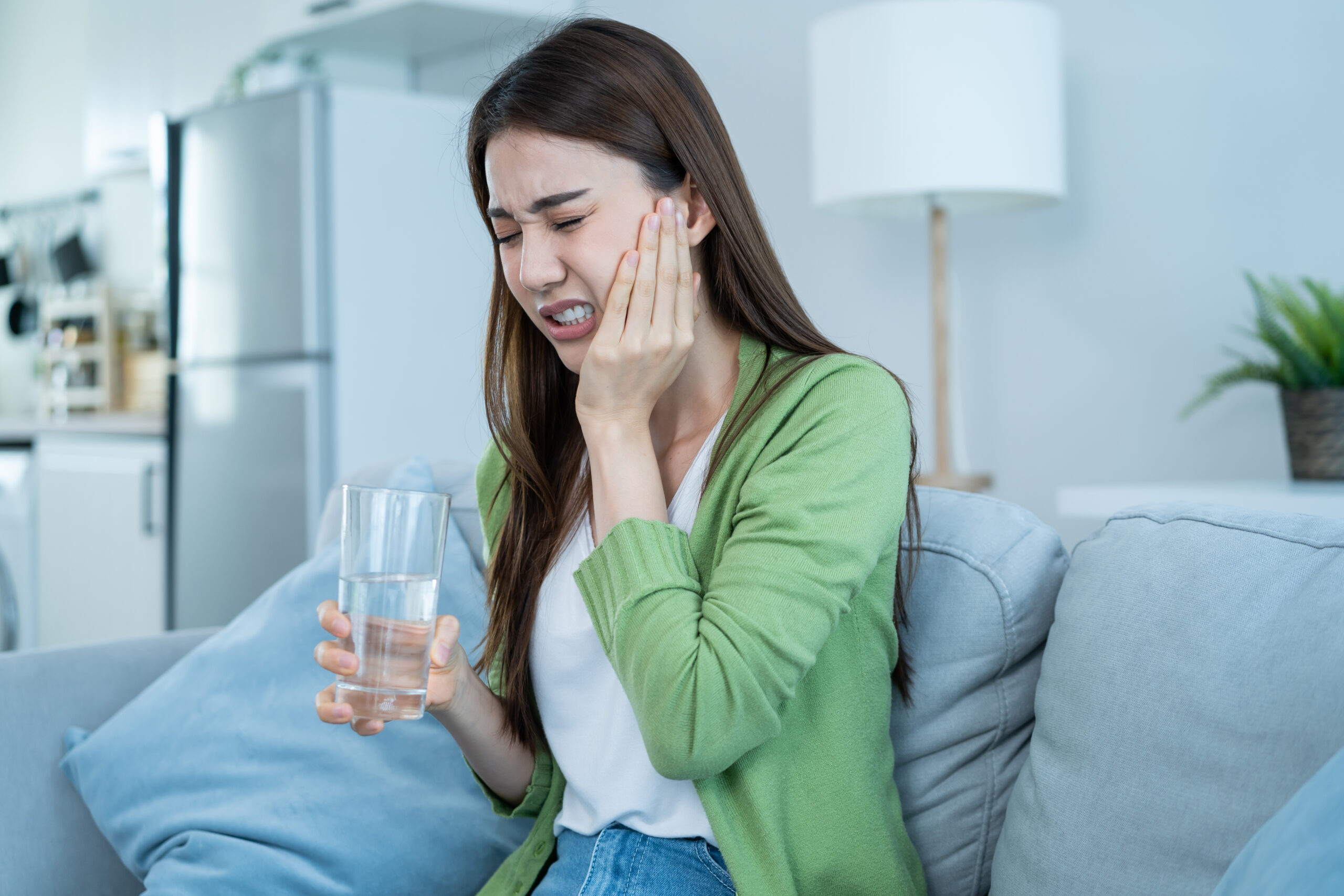 Woman experiencing severe tooth pain while holding a glass of water at home, seeking help from an emergency dentist in Peoria.